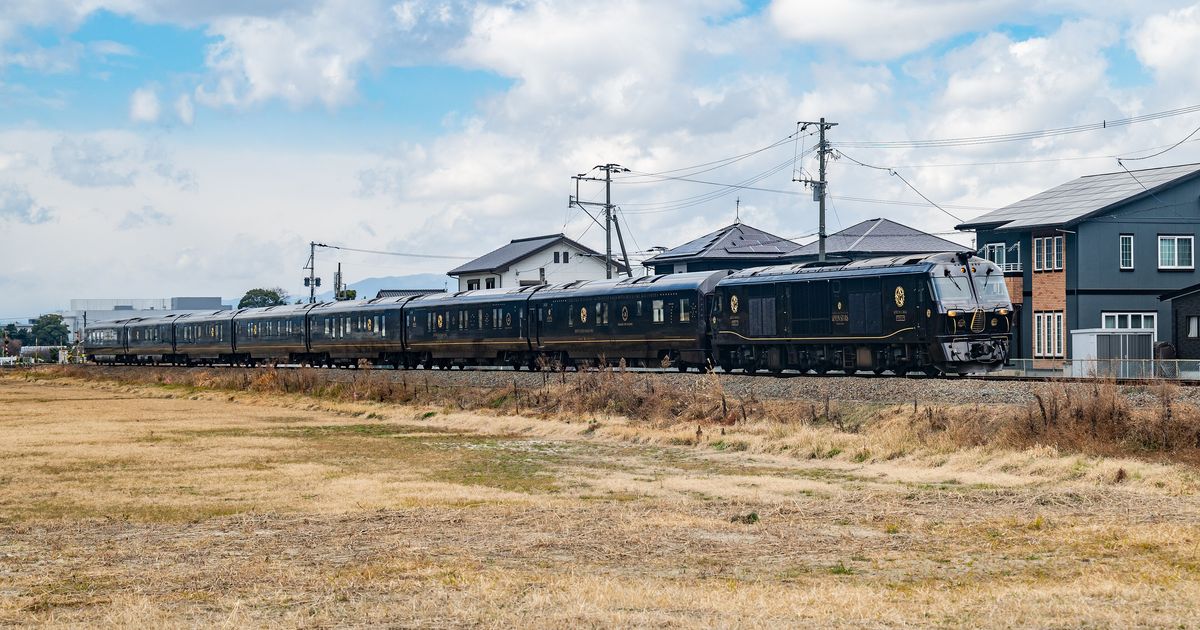 九州七星號整列行駛在住宅旁的鐵道上:最前面是深色車身、金色徽章的柴油機車,後面拖著 7 節同色客車,前景是冬天枯黃的田地,背景是藍天白雲