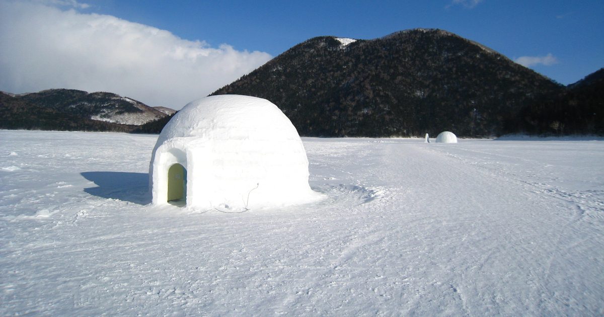 結冰的然別湖湖面上一棟雪塊砌成的圓頂冰屋，入口朝左，遠處湖面還有另一棟小冰屋，背後是覆著樹林的天望山與藍天