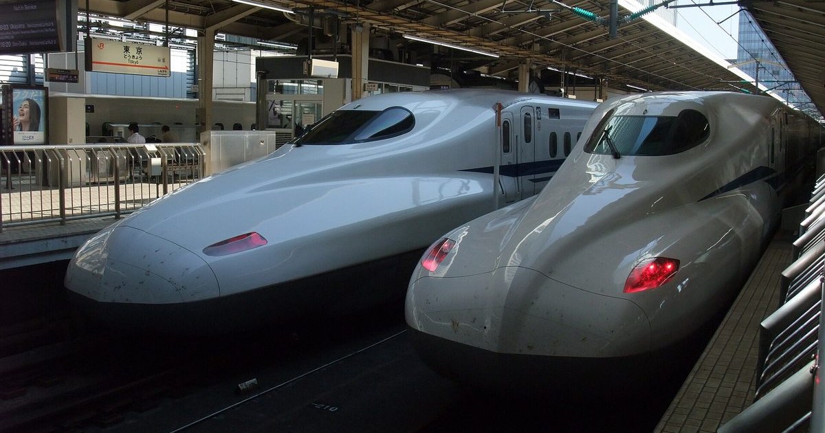 Two white Shinkansen trainsets nose to nose at a Tokyo Station platform, duckbill noses and blue bodyline stripes, the right-hand set showing red tail lights; the JR “Tokyo“ platform sign and a departure board are visible at upper left