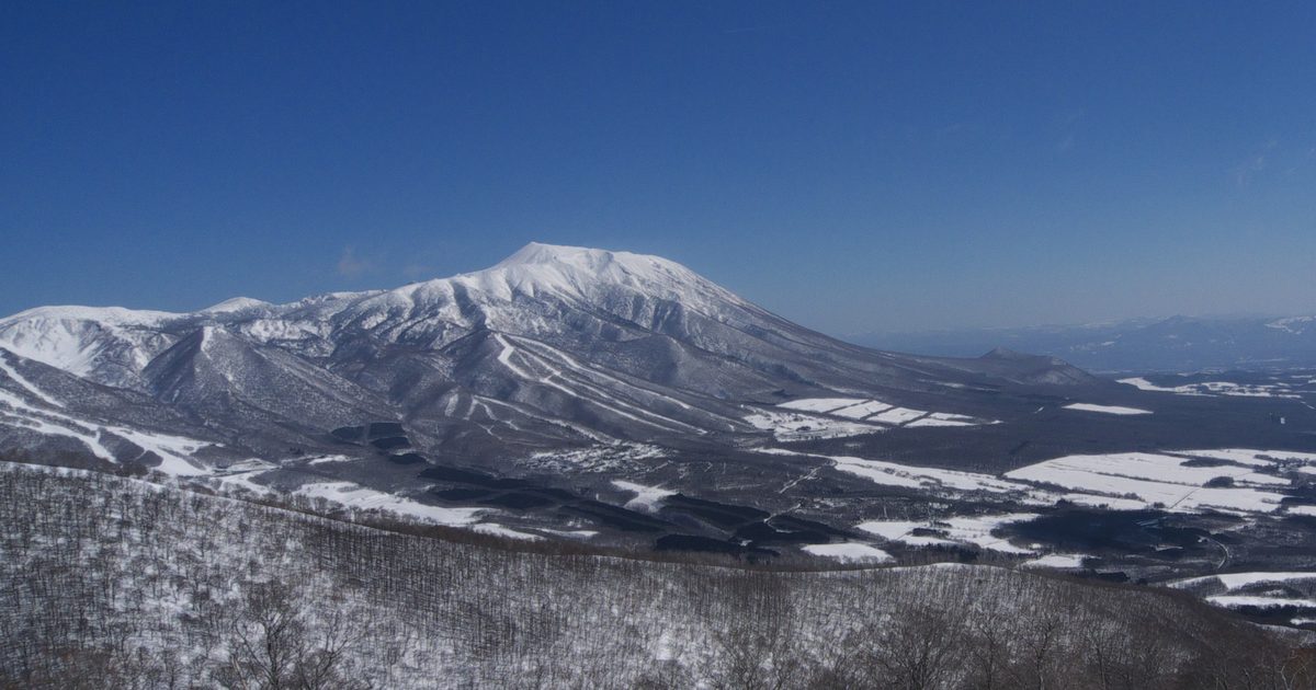 3 月晴天從雫石滑雪場高處望出去:藍天下積雪的岩手山與鞍掛山,山麓是黑色樹林與白色雪原,前景是覆雪的落葉林斜坡