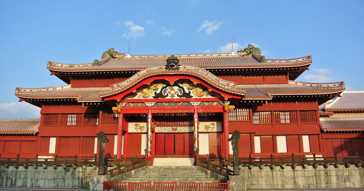 The Seiden of Shuri Castle seen head-on: a vermilion-lacquered timber hall with a double roof, a gilded dragon-and-peony carving under the central curved gable, a green dragon-head ornament at each end of the ridge, a carved grey stone dragon pillar standing on either side of the front steps, and a coral-limestone platform below (photographed February 2015 — the Heisei-era reconstruction that burned down in 2019)