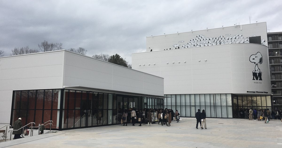 南町田グランベリーパーク的史努比博物館一帶:右邊是白色方形的博物館本館,圓弧外牆上畫著坐在 M 字上的史努比與 TOKYO 字樣,頂樓外牆有一整排史努比圖案;左邊是較低的白色玻璃帷幕建築,前面是寬闊的石板廣場與排隊的遊客,陰天