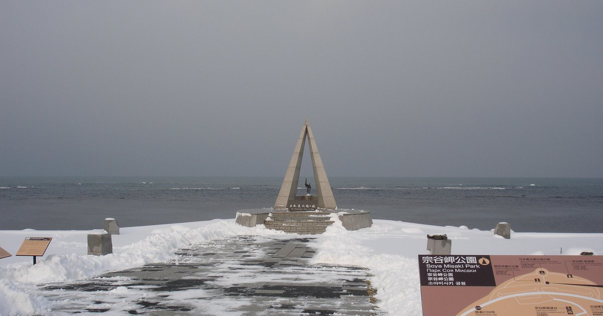 12 月的宗谷岬:剷過雪的石板步道通往三角形的「日本最北端の地」碑,碑後是灰色的海與陰天,步道兩側積雪,右下角是宗谷岬公園的導覽看板