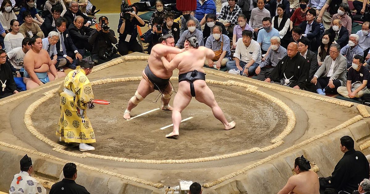 Two sumo wrestlers locked on to each other's belts at the centre of the clay ring while a gyoji in a yellow robe holds up a red gunbai fan, with spectators seated on cushions all the way around the ring