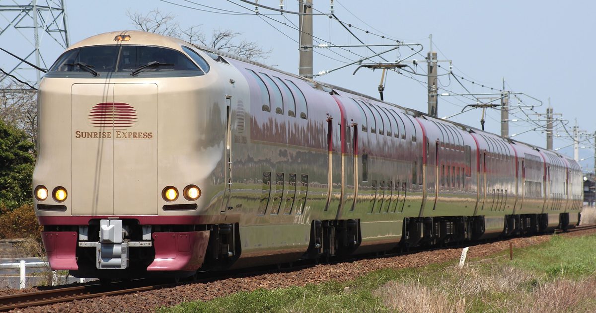 晴天鄉間的電氣化鐵道上,一列 285 系 Sunrise 列車迎面駛來:米白色車頭印著紅色橫條太陽標誌與「SUNRISE EXPRESS」字樣,車燈亮著,後方一長串雙層車廂上半紅、下半米白,車身反射著草地,前景是枯黃的草