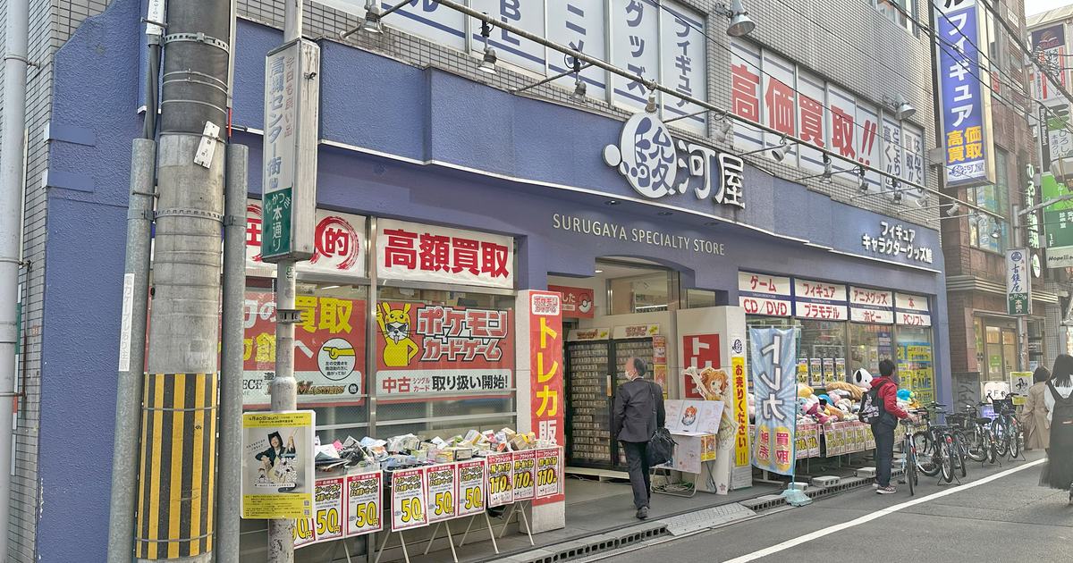A Suruga-ya storefront with its blue-and-white sign, red buy-back banners and trading-card notices