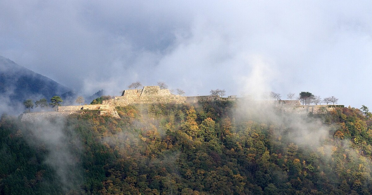 Takeda Castle ruins seen from the mountain opposite: bare stone ramparts strung along the ridge with mist drifting across the slope and autumn colour in the forest below
