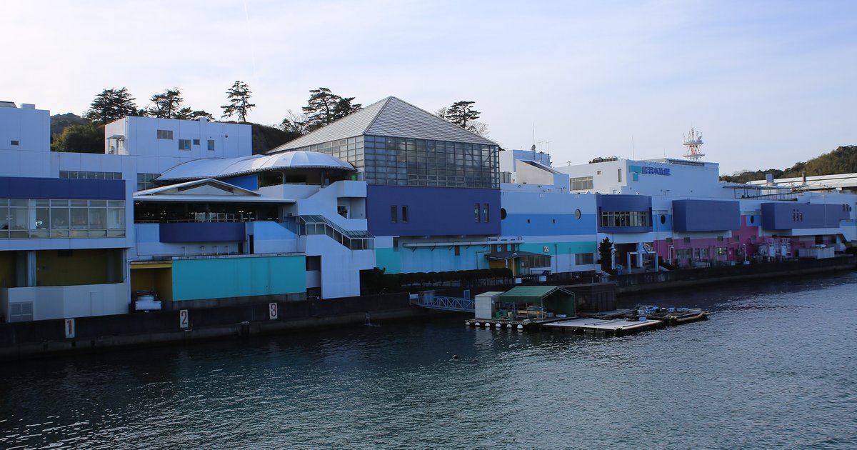 從鳥羽港海面看鳥羽水族館:沿岸一整排藍、白、水藍與粉紅色塊的館舍,中間是玻璃斜屋頂的展館,岸邊停著小浮台