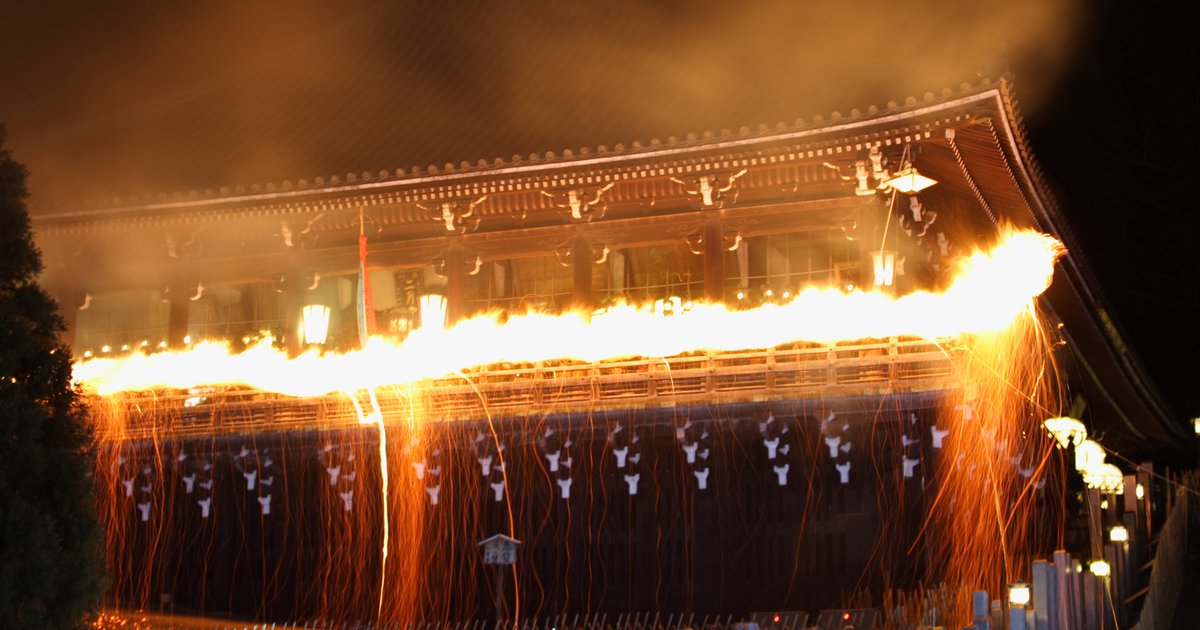 夜裡從下方仰望東大寺二月堂:松明沿著舞台欄杆移動,火焰在欄杆上拉成一條火帶,火粉像雨一樣往下灑,屋簷下掛著燈籠,上方被煙燻成橘色