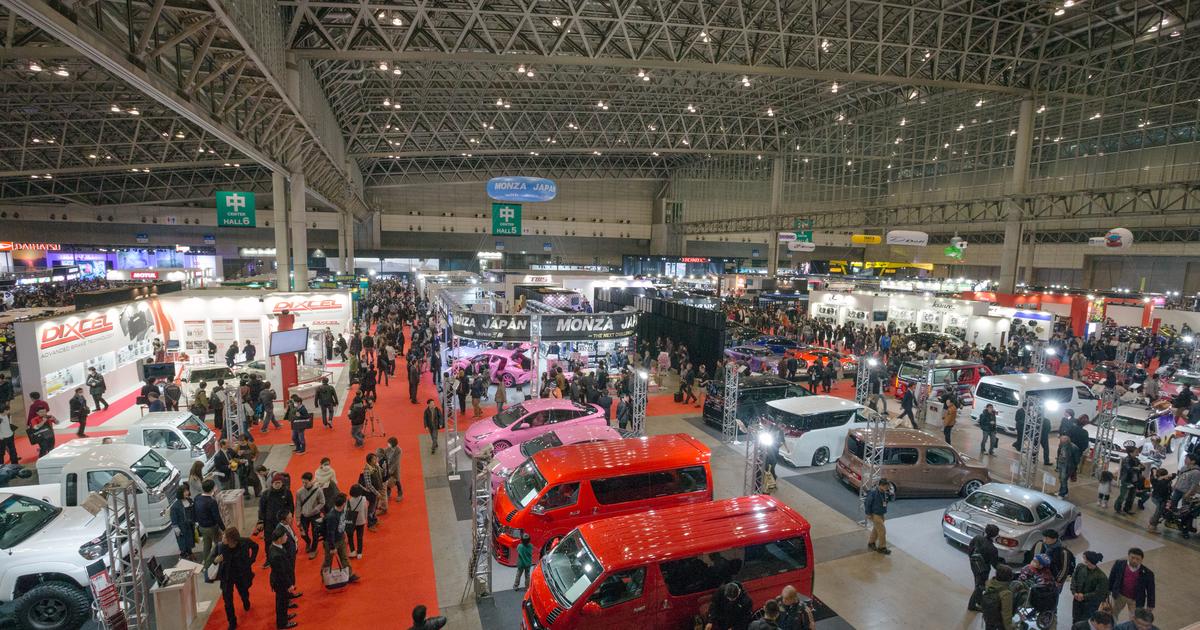 從高處俯瞰東京改裝車展的展館:鋼骨格狀天花板下,紅地毯走道兩側排滿改裝車攤位,前方是兩台紅色廂型車與粉紅色轎車,遠處掛著綠色「中 HALL 5」「HALL 6」展館標示,走道上人潮不斷