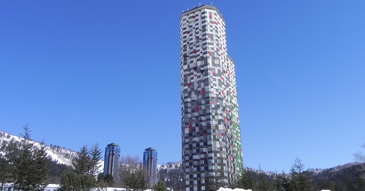 Hoshino Resorts Tomamu on a clear winter day, the mosaic-clad Tower rising in the centre with two blue glass towers behind to the left and a row of small conifers on the snow in front