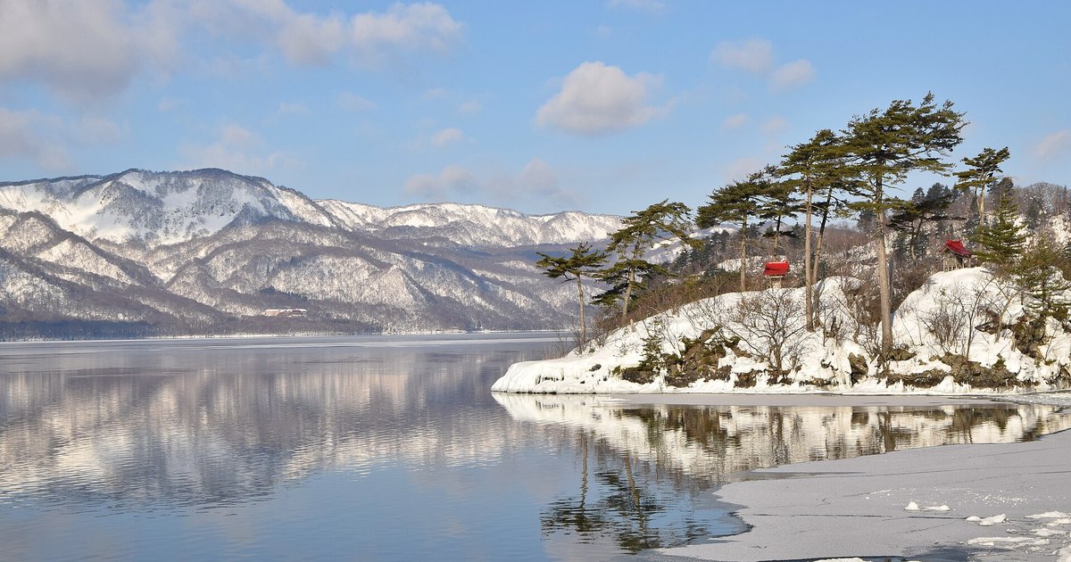 2022 年 2 月的十和田湖休屋湖畔：右邊是積雪的小島恵比寿大黒島，島上幾棵赤松與兩座紅色屋頂的小祠，左邊是對岸覆雪的山，平靜的湖面映著山與雲，右下角的岸邊結了一層薄冰