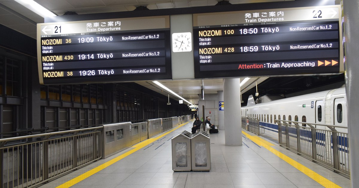 A Shinkansen stopped at a Shinagawa Station platform, car numbers marked beside the doors, with overhead departure boards listing Nozomi services and their non-reserved cars