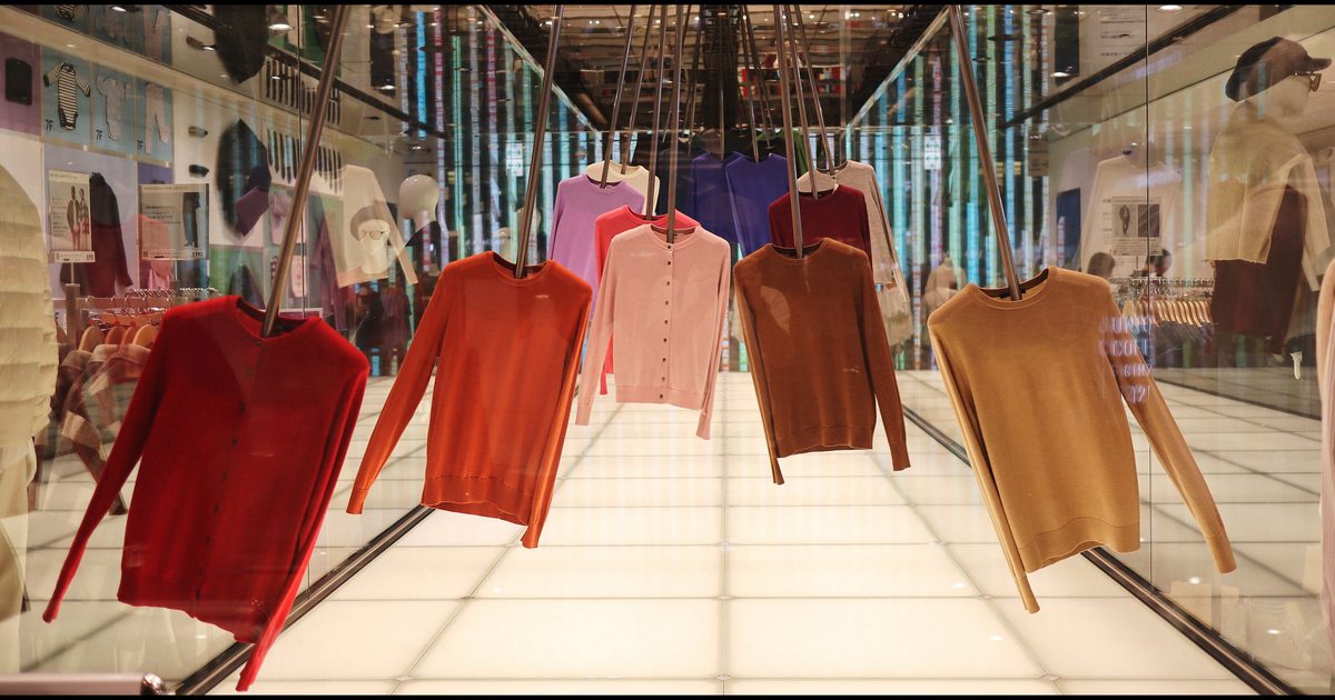 UNIQLO 銀座店內的展示：一排紅、橘、粉、棕、芥末黃的針織衫用衣架懸吊在發光的白色地板上方，兩側玻璃後面是掛滿衣服的衣架，2023 年 9 月