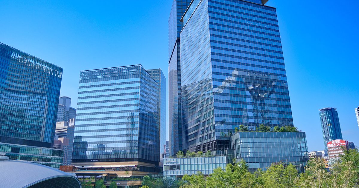 The Umekita district north of Osaka Station on a cloudless day: newly planted trees and low greenery in front, three glass-clad towers behind, the tallest capped by a projecting roof canopy, and a red rooftop sign for the Shin-Umeda training centre in the distance to the right