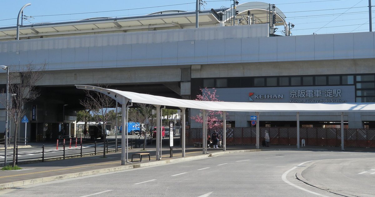 晴天的京阪淀站高架站舍,牆上寫著「京阪電車 淀駅」,站前公車候車棚旁一棵開滿粉紅花的早櫻