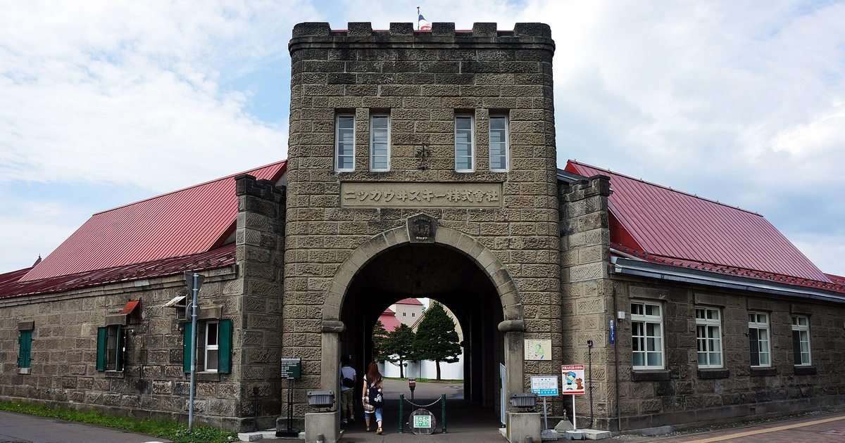 余市蒸餾所正門:灰褐色石砌的城垛塔樓,門額刻著「ニッカウヰスキー株式會社」,下方是拱門,兩側延伸出紅色屋頂的石造建築