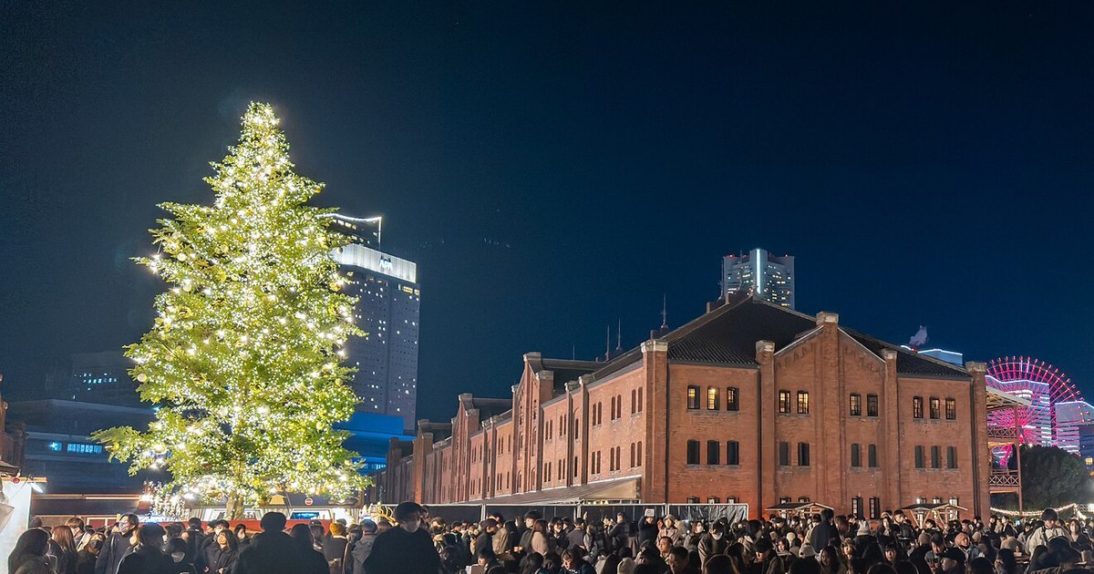 2022 年 12 月橫濱紅磚倉庫聖誕市集的夜景：左邊是一棵纏滿暖白色燈串的大聖誕樹，右邊是紅磚倉庫的山牆與幾扇亮著燈的窗，倉庫後方露出粉紅色的摩天輪，畫面最下緣是廣場上滿滿的人群