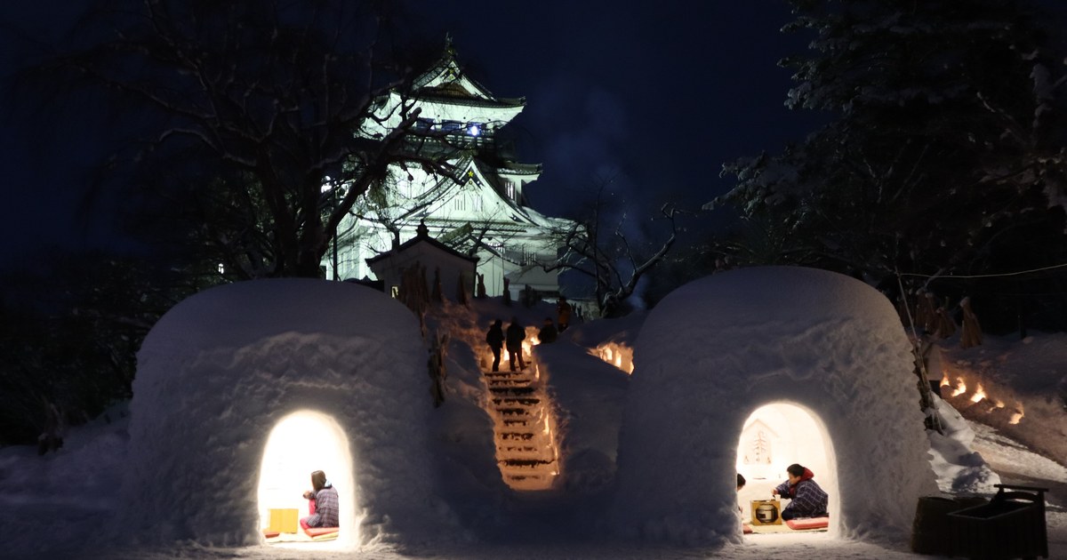 Two snow domes glowing from within at Yokote Park at night, children sitting inside each, a candle-lined snow staircase between them and the floodlit Yokote Castle keep behind