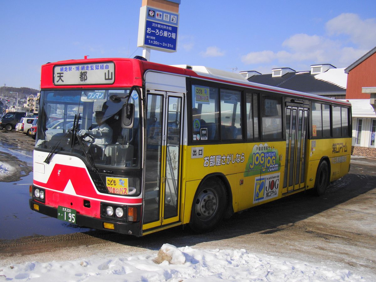 停在雪地停車場的紅白網走巴士,車頭行先牌寫「網走駅・(博)網走監獄経由 天都山」,後方藍色看板寫「流氷観光砕氷船 おーろら乗り場 運行期間 1月20日〜3月31日」