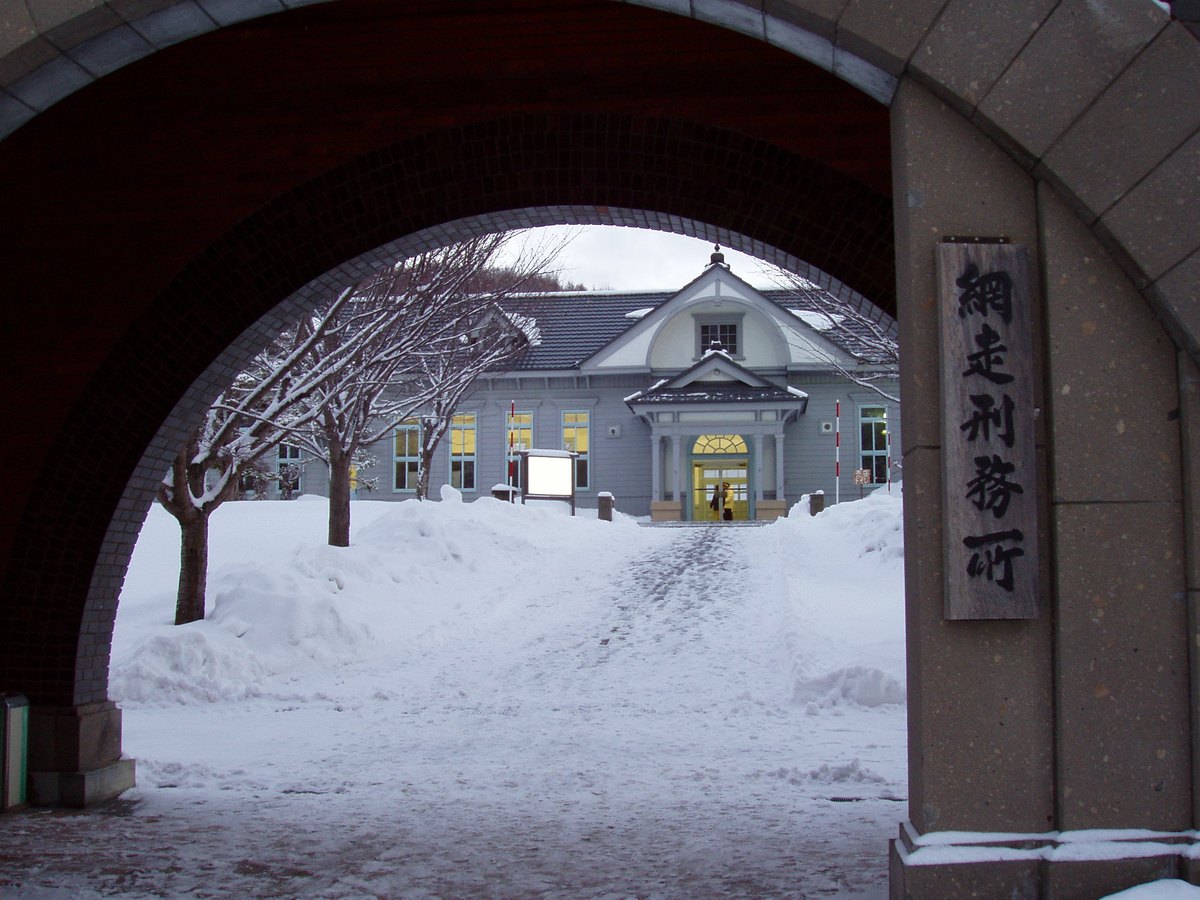 從紅磚正門的拱門下往裡看:右邊石柱掛著直書「網走刑務所」的木牌,雪地小路盡頭是一棟淺灰藍色的木造洋風建築,窗裡亮著燈