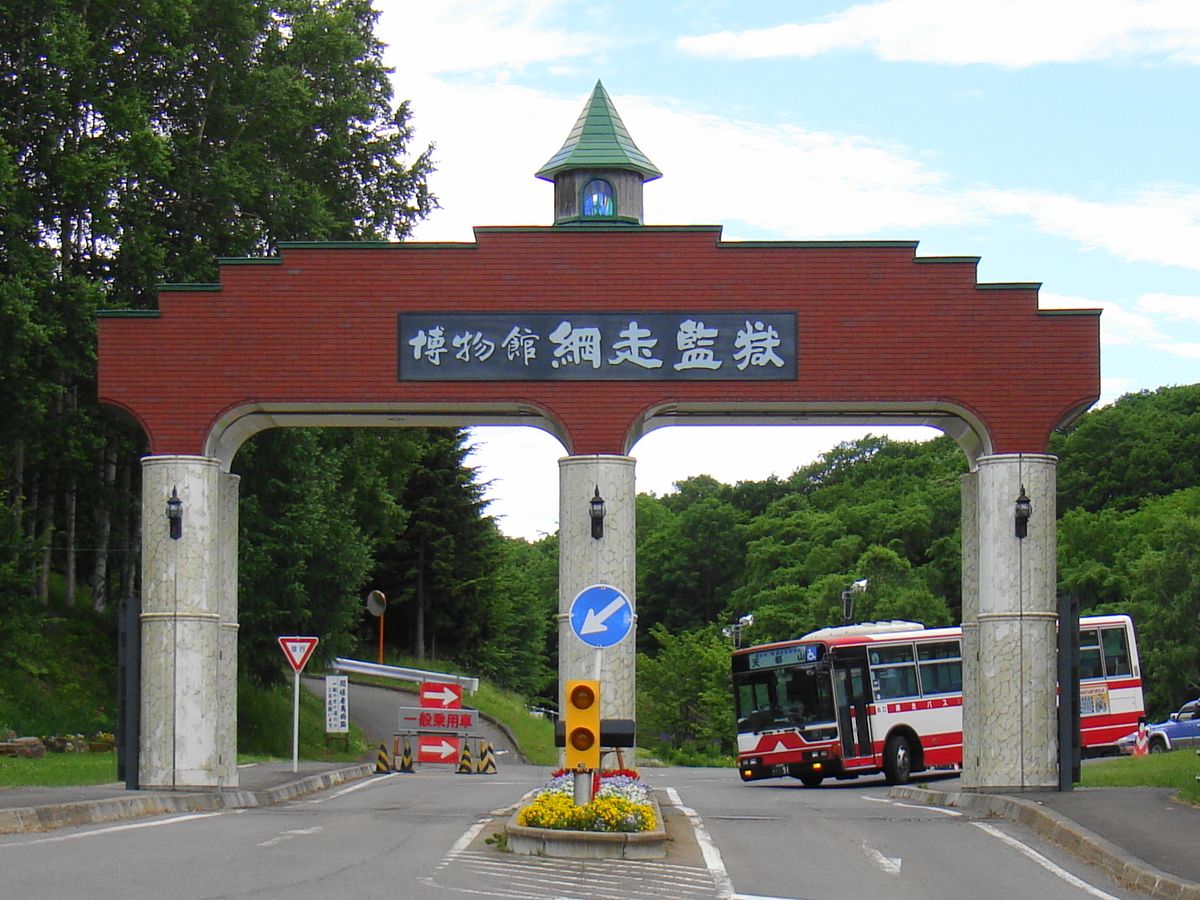 紅磚色的大拱門,正中寫著「博物館 網走監獄」,拱門下是進館的道路,路的後方有一輛紅白網走巴士