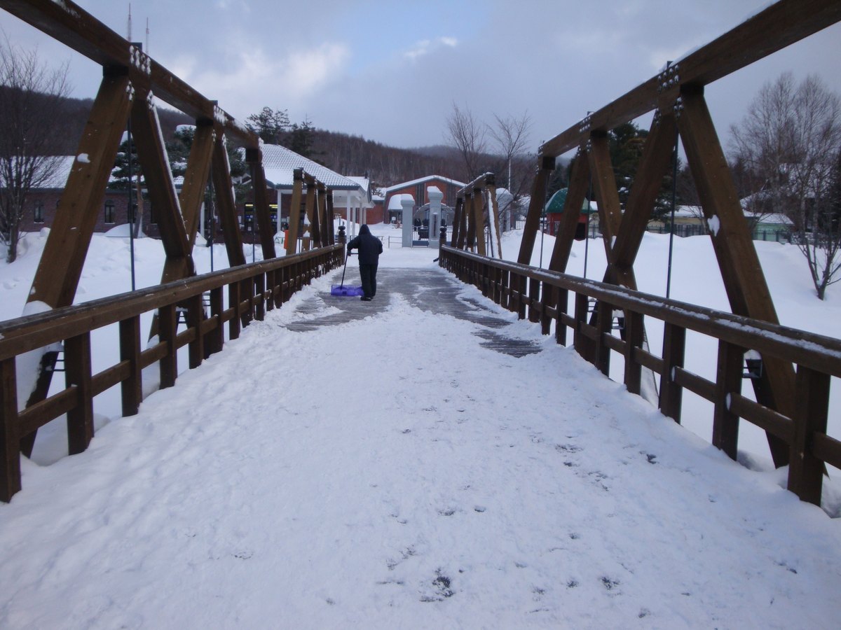 積雪的木造桁架橋,橋面中間剷出一條路,遠處一個人背對鏡頭在推鏟雪工具,橋的另一頭是入口一帶的建築