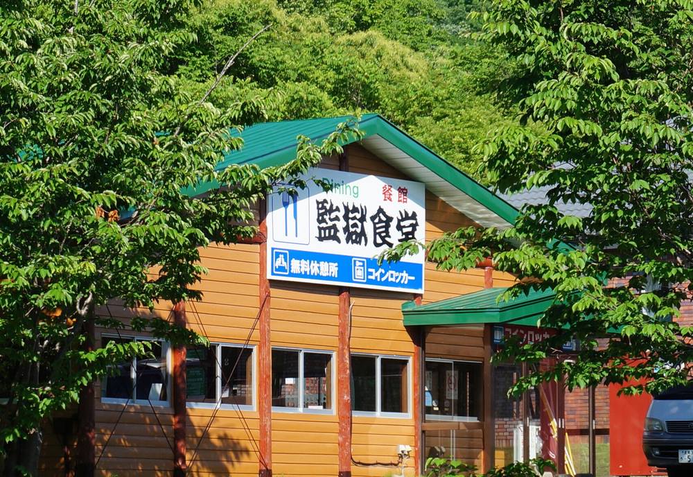綠色屋頂的木造平房,牆上白底招牌寫「監獄食堂」,招牌下方藍底小字寫「無料休憩所」與「コインロッカー」,前面有樹遮住一部分