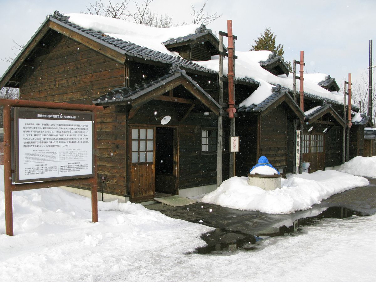 雪還在下的木造平房,屋頂堆著厚厚一層雪,門口前立著一塊解說牌,地面積雪