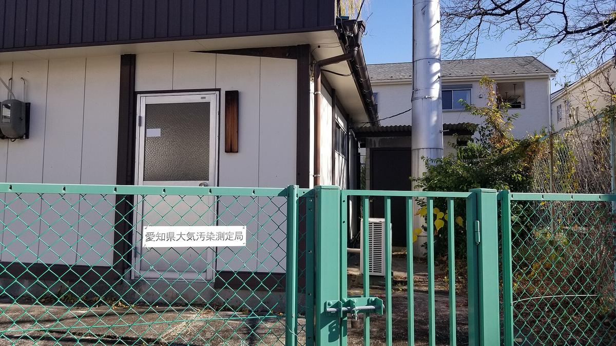 A small white single-storey hut in a residential street, a plate beside the door reading 愛知県大気汚染測定局, a stainless steel sampling stack rising above the roof, and the whole plot ringed by a green wire fence with a padlocked gate