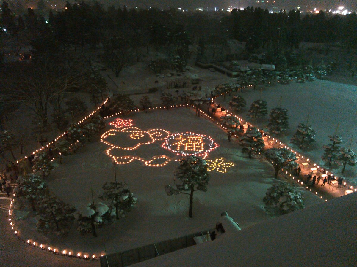 從天守閣往下看的積雪廣場：地上用燭火排出一個卡通人物和圓圈裡的「福」字，旁邊一排黃色燭火，右側步道兩旁排著燈籠與燭火，步道上有小小的人影