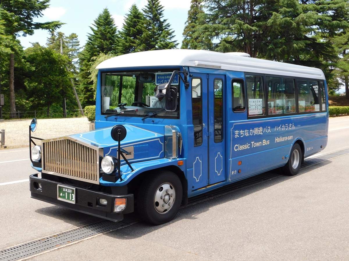 藍色車身、前面有復古金屬水箱罩的小型巴士，車側寫著「まちなか周遊バス ハイカラさん」「Classic Town Bus」，停在綠樹旁的路邊