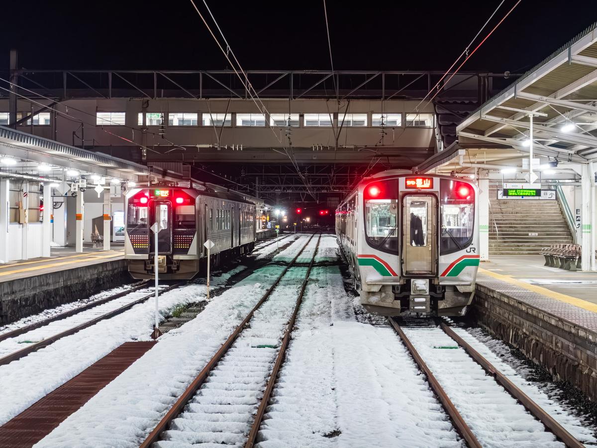 2025 年 12 月夜晚的會津若松站:軌道上積著一層雪,兩列亮著紅色尾燈的列車停在月台兩側,右邊那列車頭顯示「郡山」,月台樓梯上方的電子看板亮著綠字