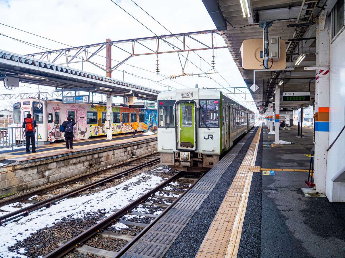2025 年 12 月的會津若松站月台:軌道間有殘雪,右側 3 號月台停著一列白底綠門、車頭有 JR 標誌的キハ110 型柴油車,對面月台掛著 4、5 號的標示牌,停著一列彩繪列車,月台上兩位旅客背對鏡頭