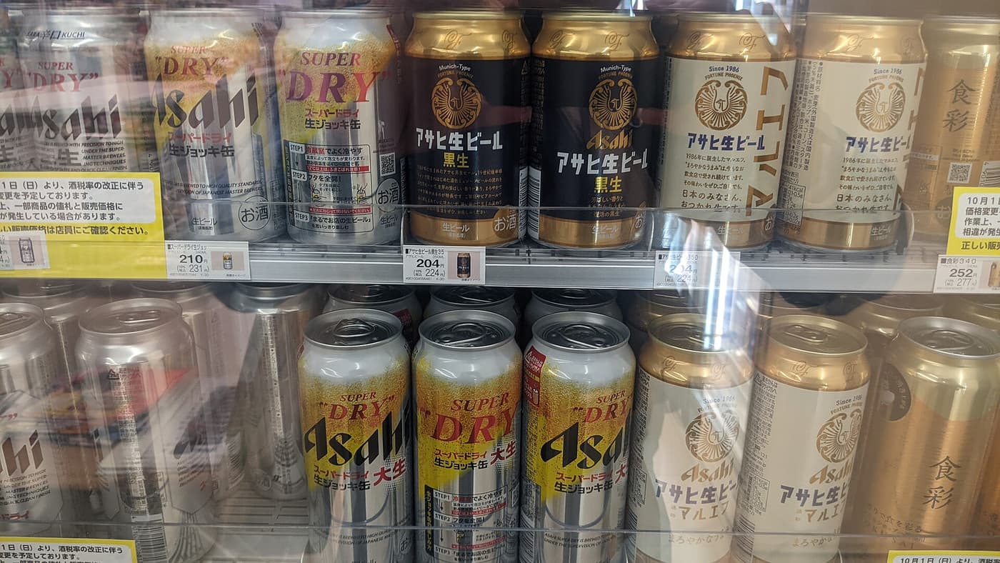 Close view inside a Japanese convenience store beer chiller: two shelves of canned Asahi beers including Super Dry Nama Jokki Kan, Asahi Nama Beer Kuronama and Maruef, white price tags clipped to the shelf edge reading 210 yen and 231 yen tax-included, and a yellow notice taped at each end of the shelf announcing a price change from October 1 due to the liquor tax revision