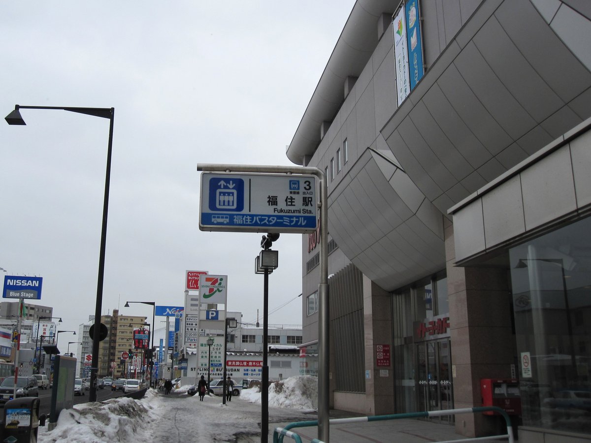 冬天的福住站 3 號出入口：大型商場建築的轉角立著藍色指示牌，寫著「3 出入口 福住駅 Fukuzumi Sta.」與「福住バスターミナル」，人行道邊堆著除雪留下的雪堆，遠處路上有幾個行人（2012 年 3 月拍攝）