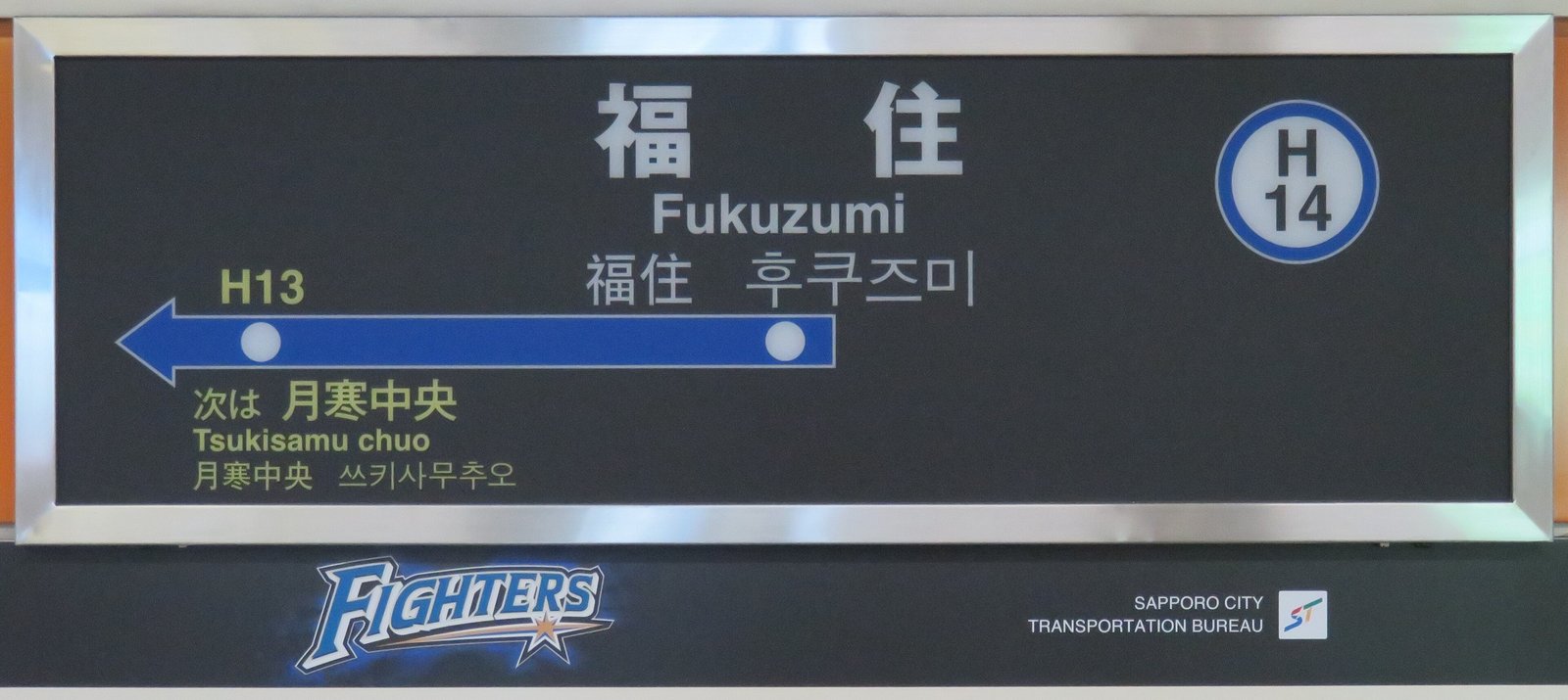 東豊線福住站月台上的站名牌：大字「福住 Fukuzumi」，右上角是藍圈車站編號 H14，藍色箭頭只往左指向下一站 H13 月寒中央，下方是北海道日本火腿鬥士隊的 FIGHTERS 標誌