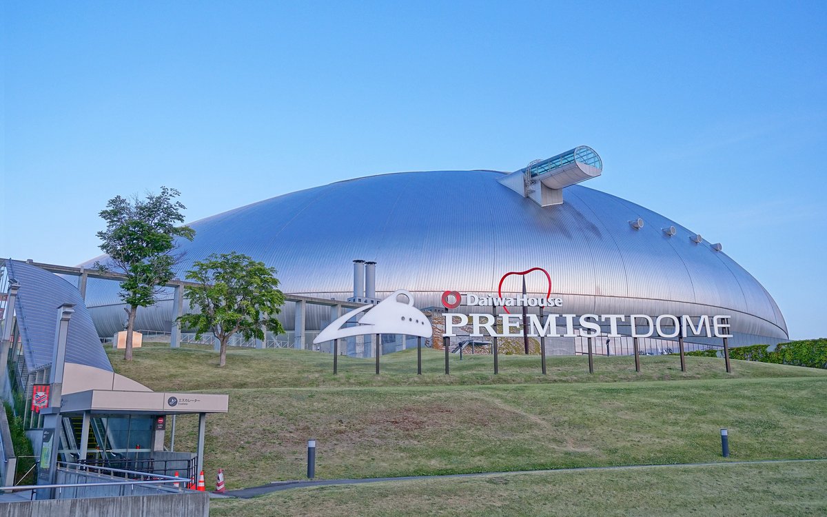 傍晚藍天下的札幌巨蛋銀色圓頂，前方草坡上立著「DaiwaHouse PREMISTDOME」的立體字招牌，左下角是通往巨蛋的手扶梯入口（2026 年 7 月拍攝，夏天）