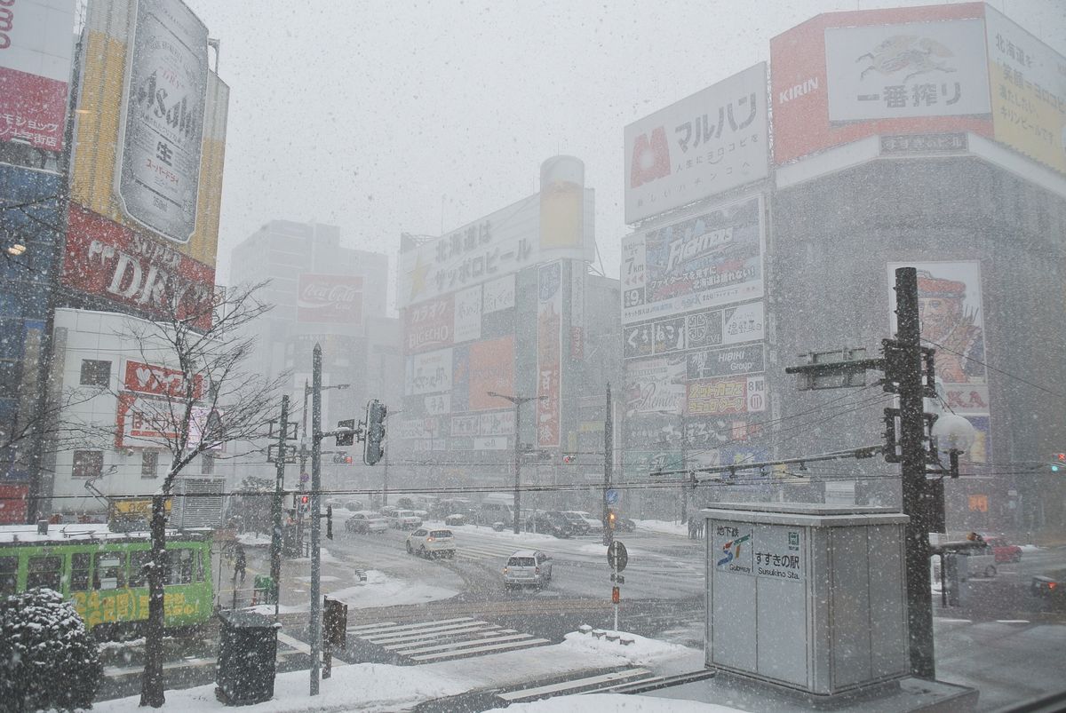 下雪中的薄野十字路口，四周大樓掛滿啤酒與威士忌的大型廣告看板，路口積雪，左下有一輛綠色路面電車，右下角是寫著「地下鉄 すすきの駅」的地下鐵出入口（2018 年 12 月 31 日拍攝）