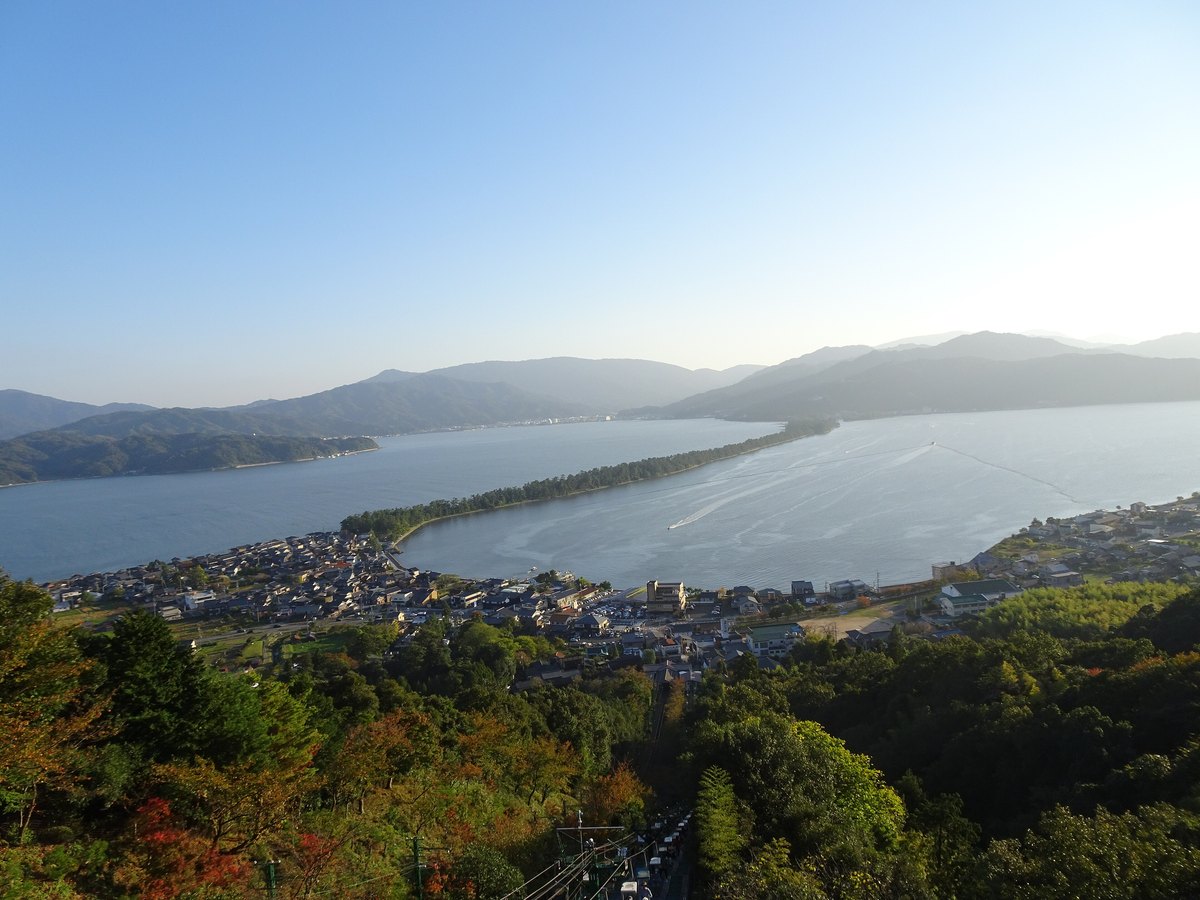 The Amanohashidate sandbar seen from a height, a thin pine-covered bar running toward the far shore, with a cable-car track cutting through the trees in the foreground