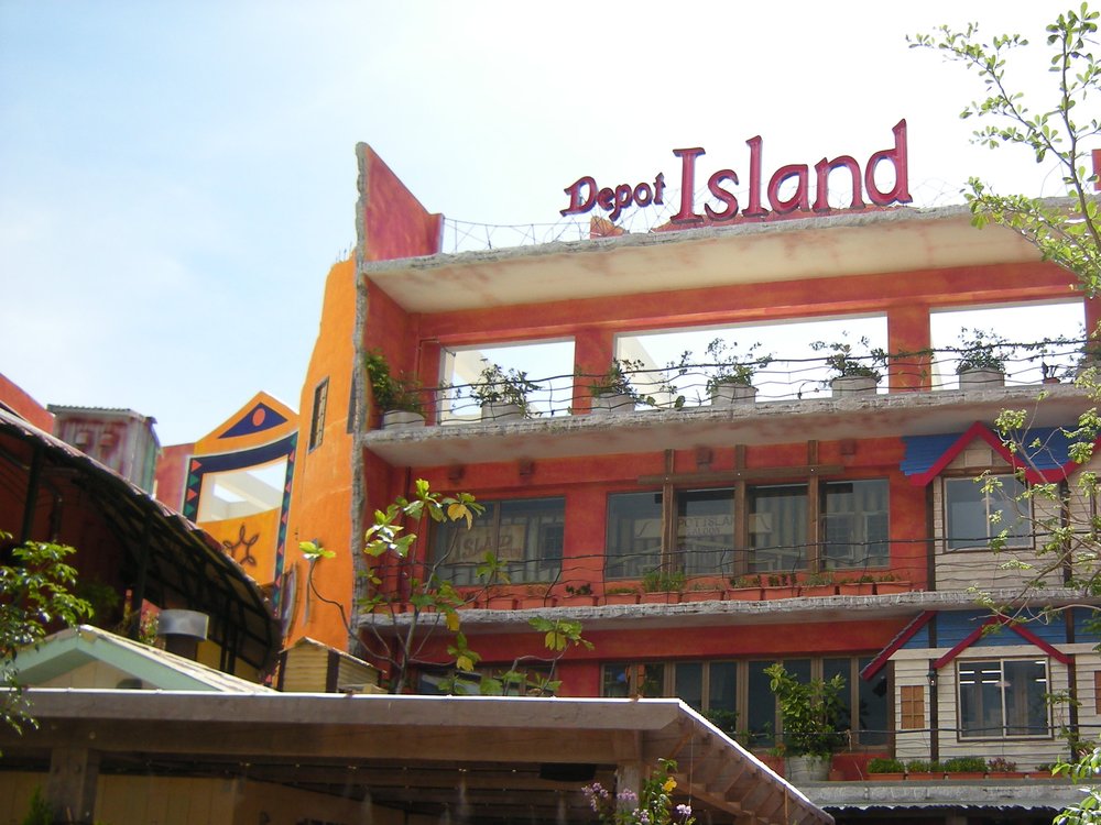 晴天白天的美國村 Depot Island 商場大樓,橘紅色外牆頂上立著紅色「Depot Island」字樣招牌,陽台擺滿盆栽,右邊有一棵細枝的樹