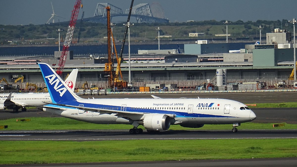 An ANA Boeing 787-8 accelerating along a runway at Haneda, blue fin, Inspiration of JAPAN along the fuselage and the registration JA878A, a Japan Airlines red crane tail visible behind it and the Tokyo Gate Bridge on the horizon
