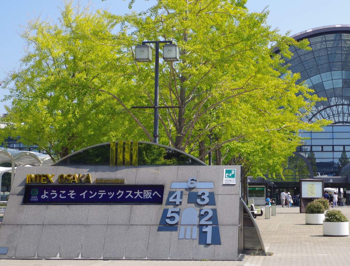 インテックス大阪西側廣場的石造導覽牌,寫著「ようこそ インテックス大阪へ」,旁邊用數字 1 到 6 標出六個展館的相對位置,右後方是玻璃圓頂與 B 號入口