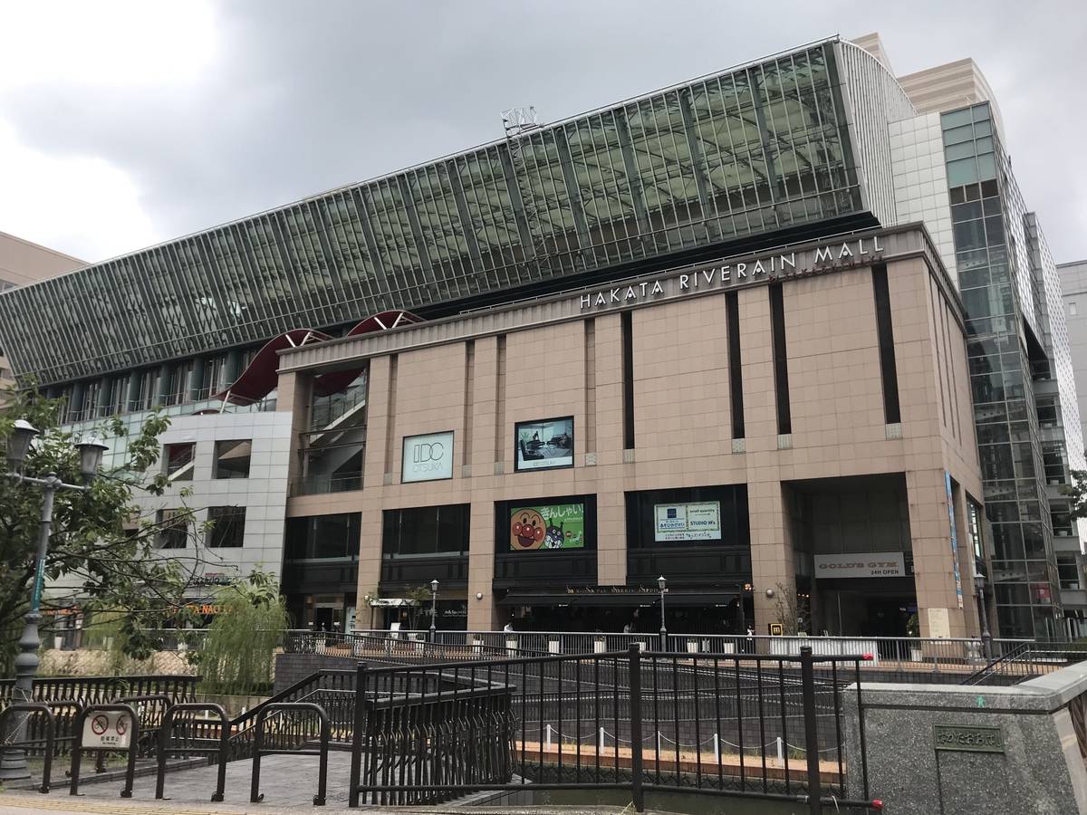 陰天從博多川邊看博多河岸商場，米色石材大樓上方寫著 HAKATA RIVERAIN MALL，頂部是斜面玻璃屋頂，前景是河邊的黑色鐵欄杆與石階