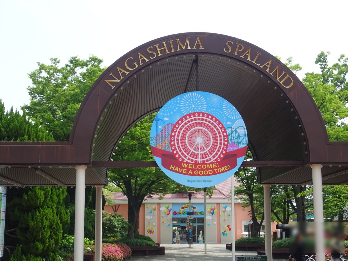 長島溫泉樂園的入口拱門，深咖啡色拱頂上寫著 NAGASHIMA SPALAND，中間掛著畫有摩天輪的圓形歡迎牌，門後是綠樹和一間粉紅色牆面的商店