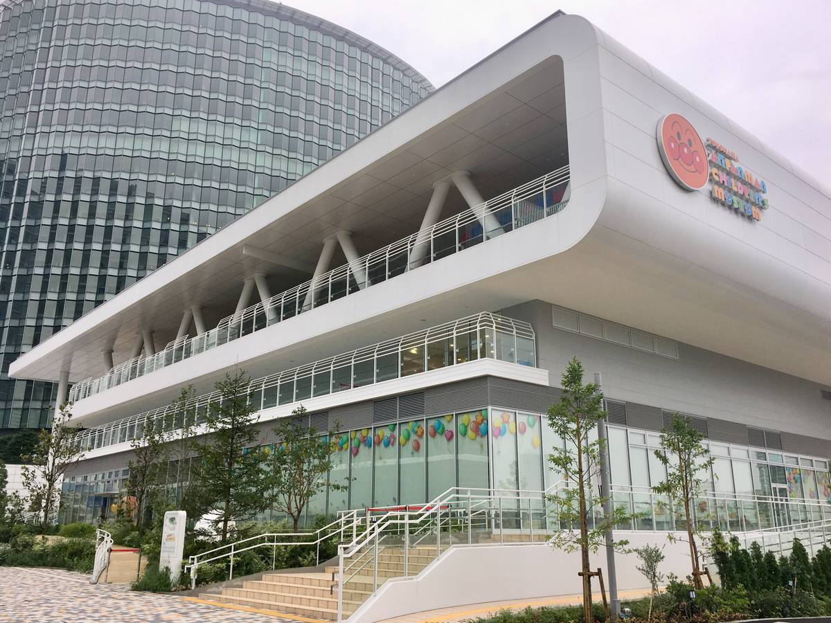 仰角看橫濱麵包超人兒童博物館的白色建築轉角，上層是外推的露台與斜撐柱，牆面有圓形麵包超人臉標誌，一樓玻璃窗貼著彩色氣球圖案，前方是通往入口的淺色階梯與扶手