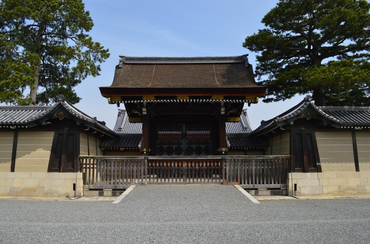 京都御所的建禮門:檜皮葺屋頂的門關著,前面一道木柵,兩側是白色橫線的築地塀,門前是一大片碎石地,左右各一棵松樹,晴天無人