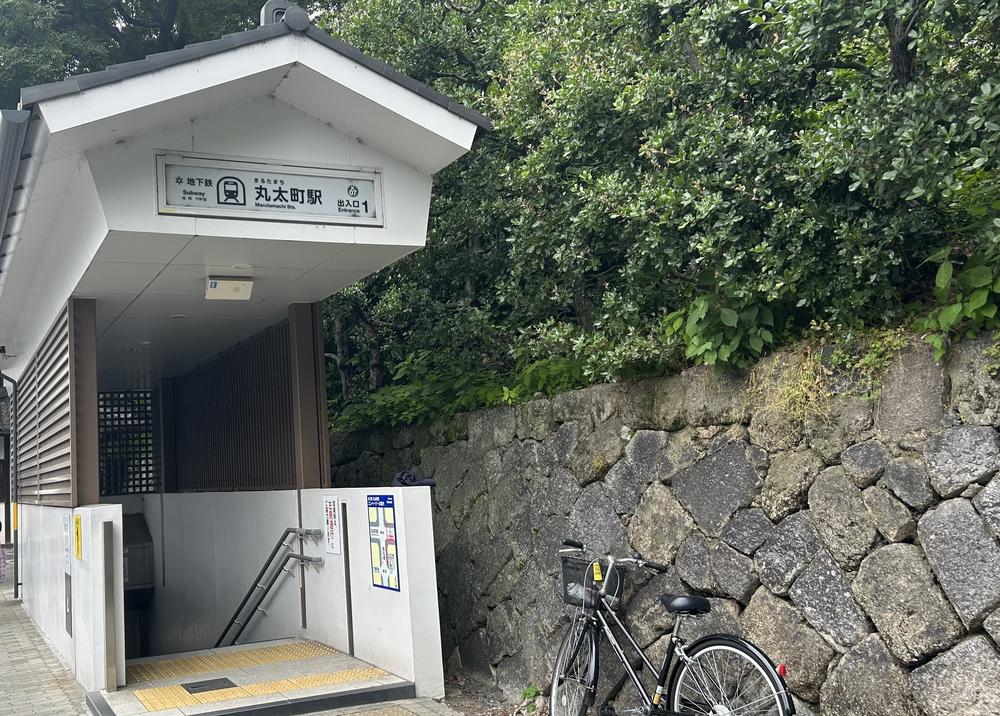 地下鐵丸太町站 1 號出口:白色牆面、灰瓦小屋頂的出入口,招牌寫著「地下鉄 丸太町駅 出入口 1」,樓梯往下,右邊是長滿綠樹的大石塊石牆,牆前停著一台腳踏車