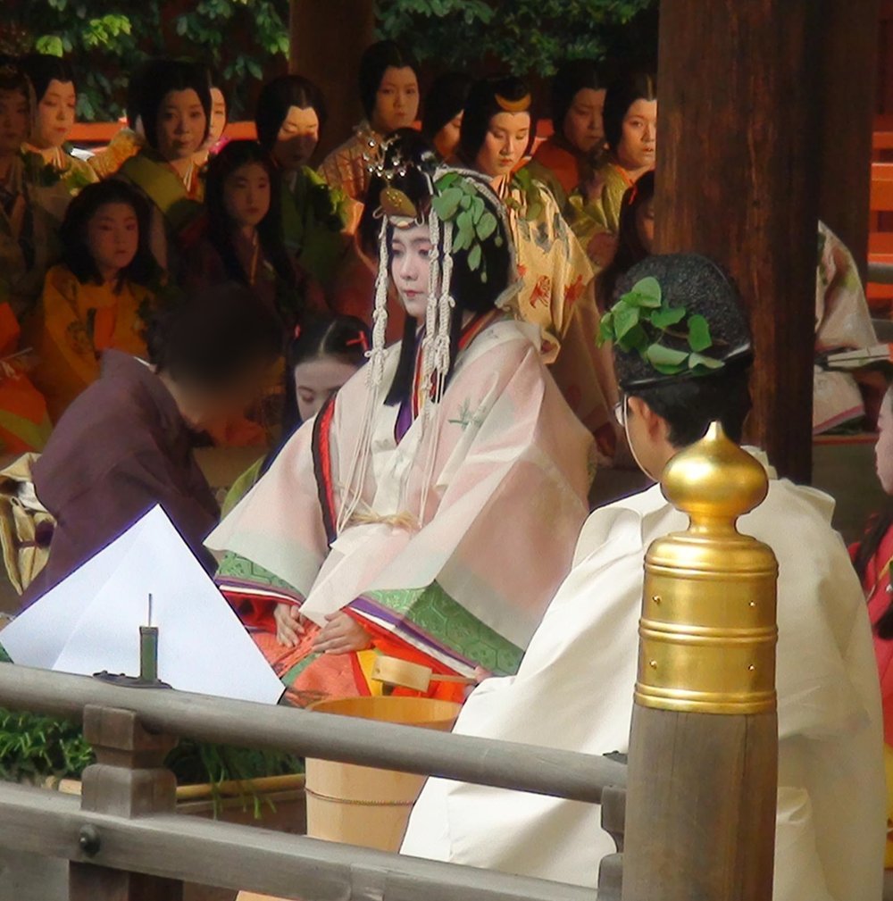 神社朱紅色迴廊下的儀式:中央一位白粉妝的女性穿淡粉色與白色的十二單、頭上垂著白色長飾並插著葵葉,面前放著木桶與白紙,前方一位戴黑冠的神職背對鏡頭,後排坐著許多穿黃、橘色裝束的女性與女孩