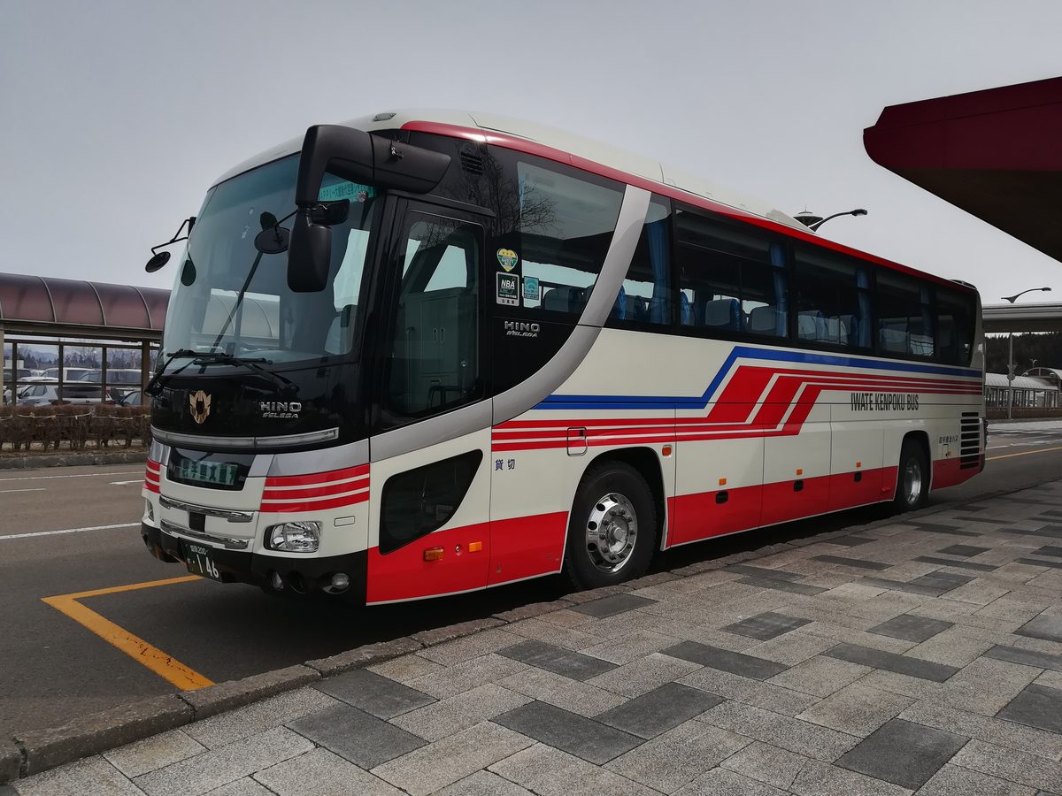 機場航廈前停著一輛白底、紅藍斜條紋的大型觀光巴士,車身寫著 IWATE KENPOKU BUS,前門旁貼著「貸切」,車頭的行先顯示看不清楚